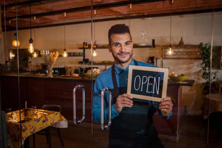 Smiling Waiter Holding Chalkboard With Open Inscription In Cafe