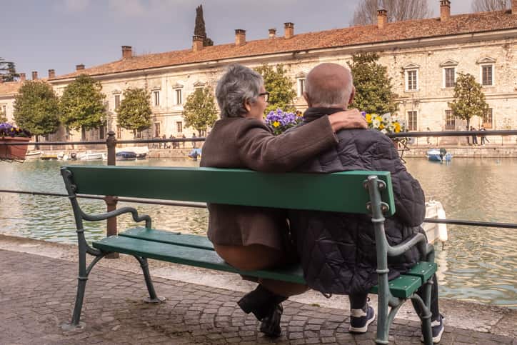 Rear view of an elderly woman embracing her husband and looking the picturesque landscape with him. A relaxing moment for an old couple sitting together on the bench in a tourist place of the Lake Garda, Italy.